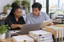 Two bookkeepers review a client intake list beside organized voucher folders.