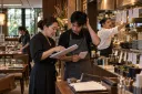 A restaurant owner reviews handwritten orders with a server as the kitchen works behind them.